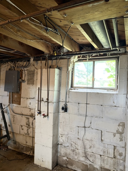 Before waterproofing and foundation repair in a Roeland Park, KS basement, showing painted concrete block walls with visible moisture staining, exposed floor joists, utility lines, and an unfinished basement window.