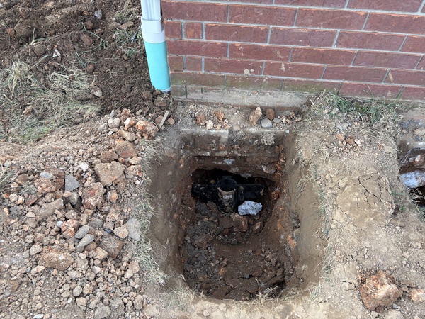 Excavated foundation area beside a brick home showing a square hole dug next to the exterior wall, exposing the footing and foundation near a downspout during foundation repair work in Cassville, MO.