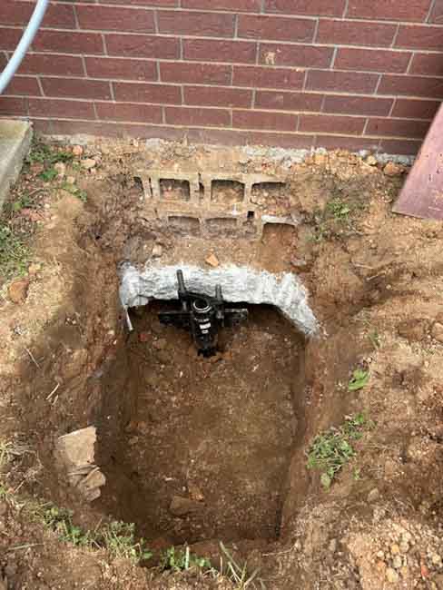 Excavation beneath a brick home's foundation showing a steel foundation pier bracket installed under the footing to provide permanent structural stabilization.
