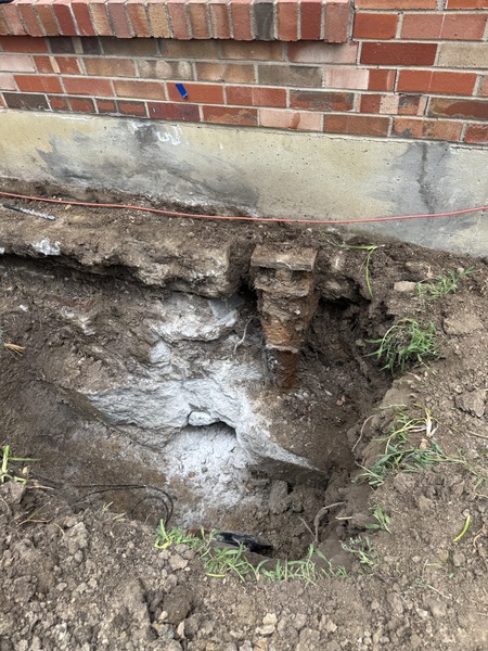 Excavation beside a brick home in Columbia, MO exposing a helical pier bracket installed beneath the foundation footing during foundation stabilization work by Spartan Ram Jack.