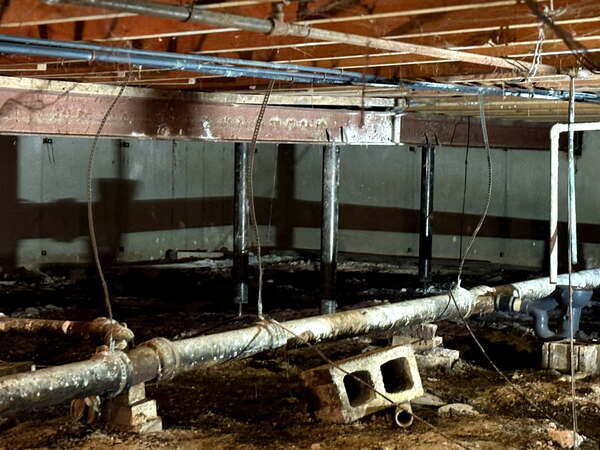 Before crawl space repair image showing temporary steel support posts, exposed plumbing, dirt floor conditions, and an uneven support beam beneath an Olathe, KS multi-family building.