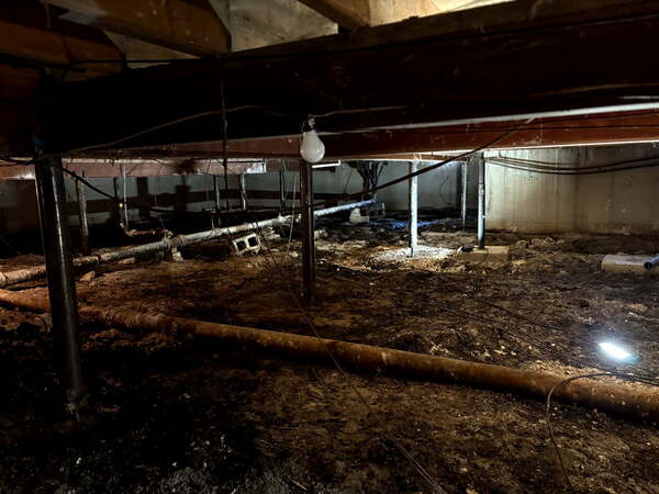 Before crawl space repair image showing a dirt-floor crawl space with sagging utilities, exposed beams, temporary support posts, and moisture-prone conditions beneath an Olathe, KS multi-family building.