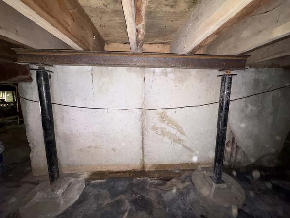 Crawl space foundation repair showing steel support posts, an overhead beam, floor joists, and a concrete foundation wall with moisture staining.