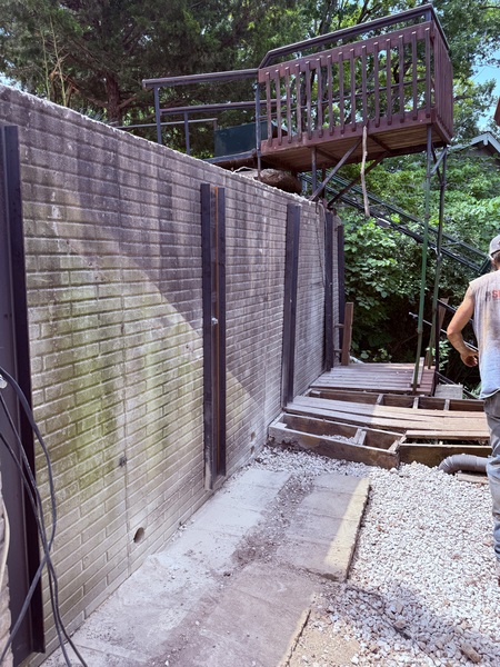 An outdoor view of a long concrete block retaining wall supported by multiple vertical dark steel earth anchors and wall brackets. A wooden deck and stairs lead up the slope, and a worker is visible on the right.