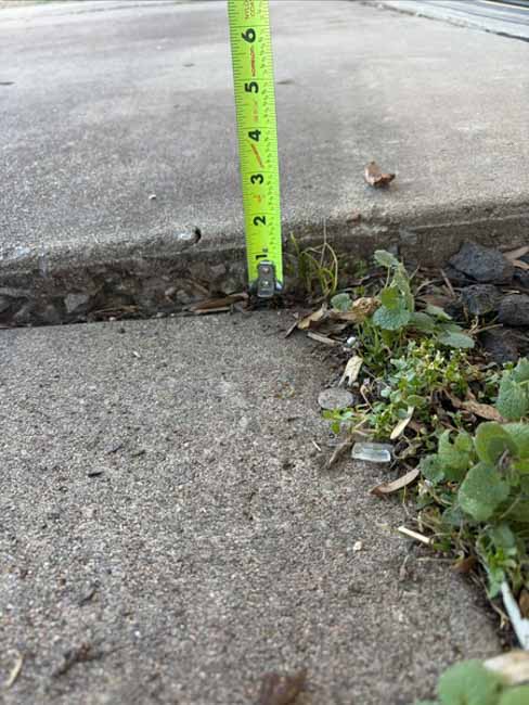 Tape measure shows approximately 5 inches of vertical separation between two concrete sidewalk slabs caused by significant settlement.
