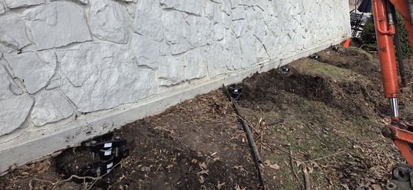 Close-up of installed black steel foundation pier brackets mounted beneath a white stone foundation wall in Wichita, KS.