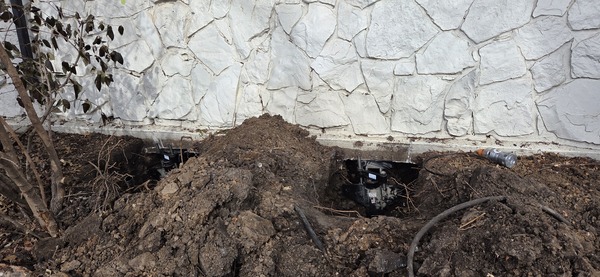 Steel foundation pier brackets installed in excavated soil pockets along a white stone residential foundation wall in Wichita, KS.